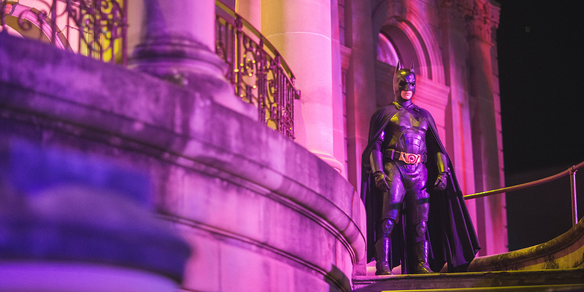 A man dressed as Batman stands on the staircase of Customs House with the evening lights glowing pink around him.