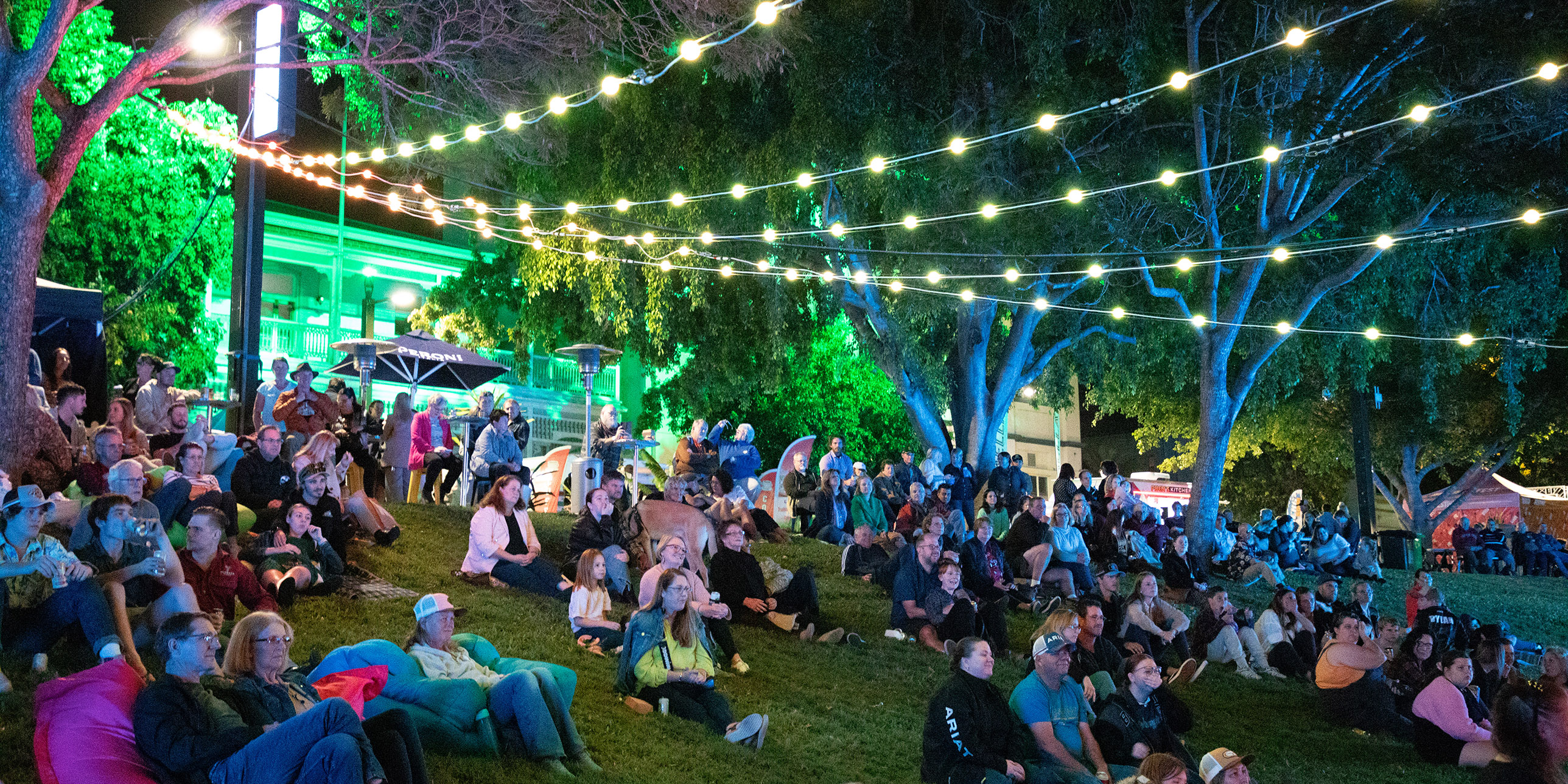 A large crowd gathers on the banks of a river enjoying the starry night and beautifully lit surrounds as live music plays in the background for the Rockhampton River Festival.