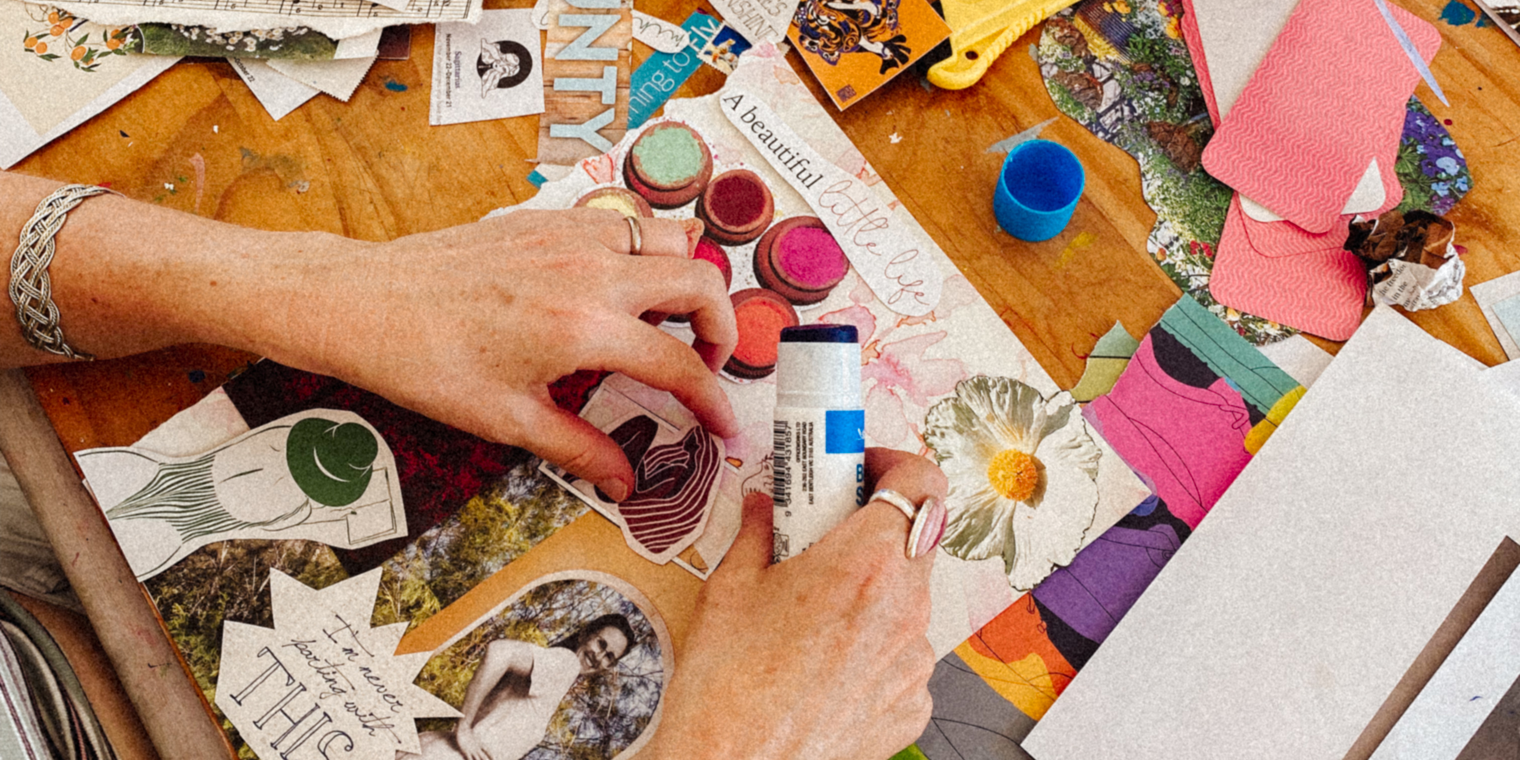 A pair of hands performing crafts on a table surrounded by paints, cut up pieces of colourful paper and drawings.