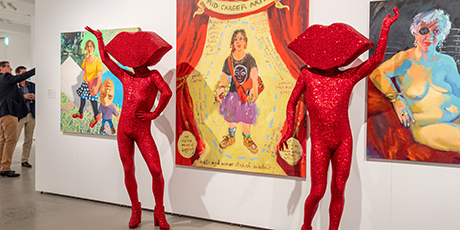 A colourful art exhibitions at Rockhampton Museum of Art featuring two people dressed completely in red body suits with lips for heads posing in front of artworks.