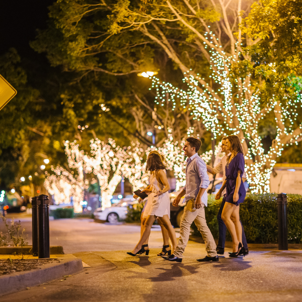 A small group of young visitors out for a night on the town. They're walking across East St among big trees full of twinkling lights.