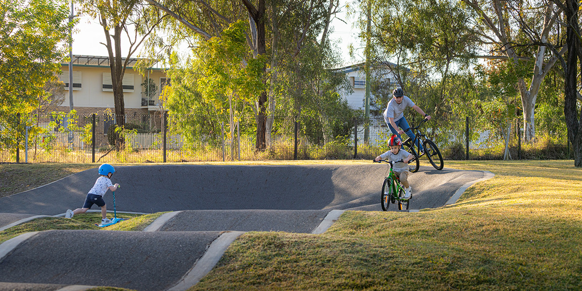 A man on a bike, a boy on a bike and a toddler on a scooter riding the pump track in morning sunlight.