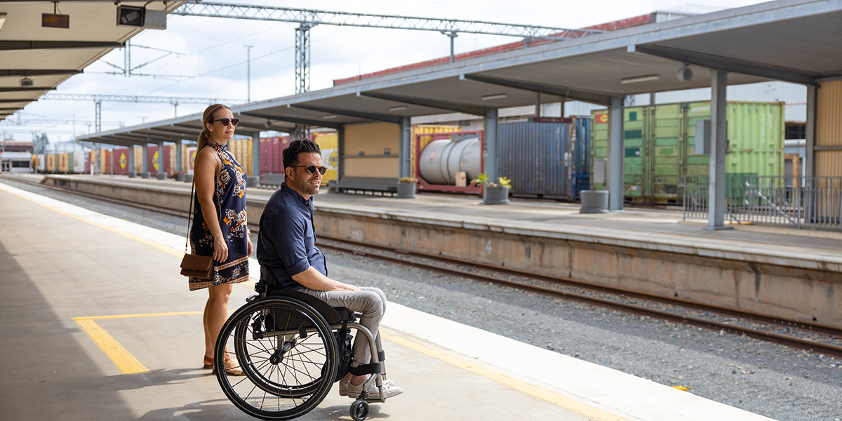 A man in a wheelchair and an able-bodied woman waiting on an a level train platform which is wheelchair accessible